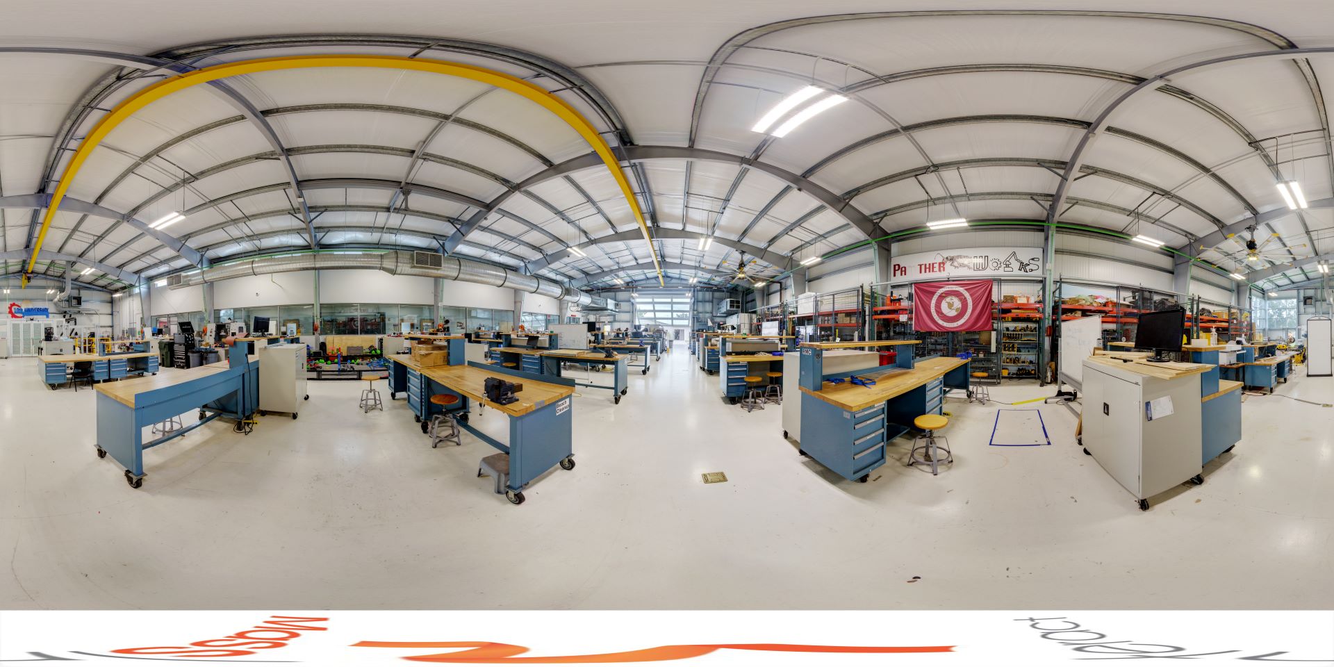Panoramic view of a spacious industrial workshop with arched metal roofs, multiple workbenches, tools, and machinery under bright lighting